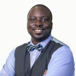 A masculine presenting person smiles at the camera their arms are crossed. They wear a blue shirt, grey waistcoat and striped bowtie